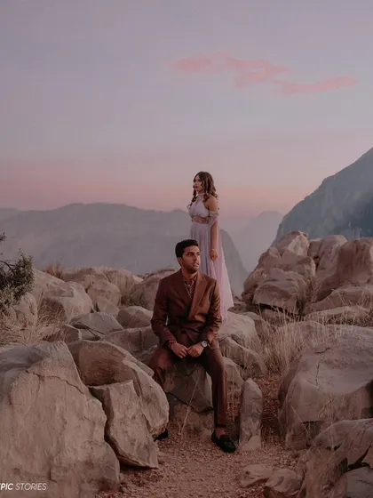 An editorial-style couple portrait set against a dramatic mountain landscape at dusk. The soft pink clouds and their poised expressions create a moody, romantic, and high-fashion feel.