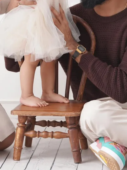A detail shot focusing on the tiny feet of a baby girl standing on a small wooden chair, held securely by her parents.