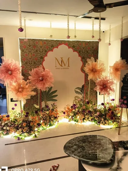 A beautiful home setup with a traditional Indian touch. The backdrop features a Mughal-inspired arch and floral print, decorated with large paper flowers and fresh flower beds.