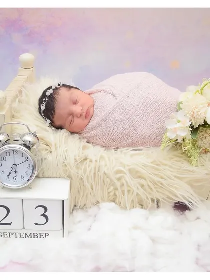A dreamy newborn setup featuring a baby sleeping on a tiny bed surrounded by fluffy clouds, with a calendar block showing a special date.