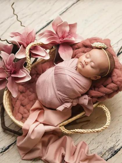 A newborn girl swaddled in pink, nestled in a basket adorned with large pink flowers. This setup is perfect for a floral-themed baby girl photoshoot.