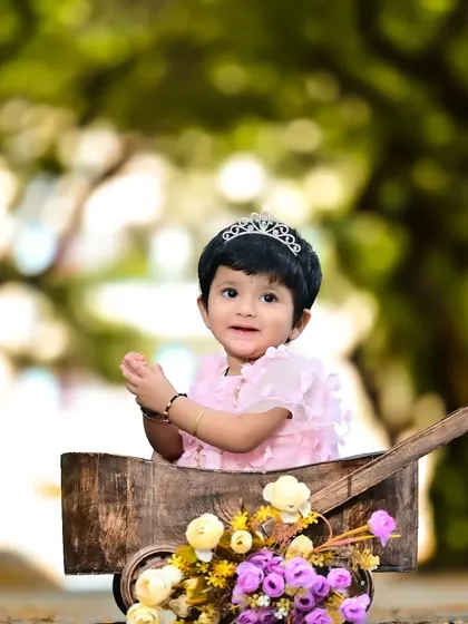 A little girl in a princess dress, sitting in a rustic wooden cart filled with flowers in a garden.