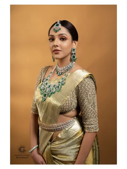 A full portrait showcasing a luxe glam South Indian bridal look. The makeup is subtle yet defining, paired with a statement emerald and diamond jewelry set against a beautiful silk saree.