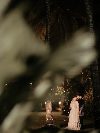 A wide shot that sets a magical scene. The couple stands together in the distance, surrounded by palm trees and fairy lights, creating a beautiful and atmospheric reception portrait.