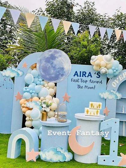 A dreamy "First Trip Around the Moon" theme for Aira's birthday. The outdoor setup features a large blue backdrop with a moon and clouds, pastel balloon garlands, and celestial props like stars and a crescent moon.
