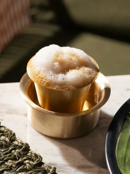 A close-up of a frothy filter coffee in a traditional brass davara. The focus is on the texture of the foam and the authentic serving style.