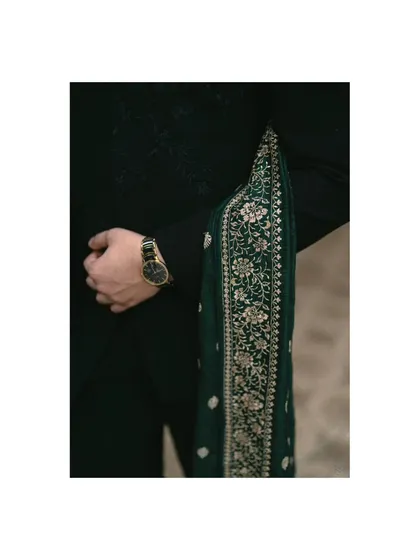 A close-up of the groom's attire, showing the rich texture of his black sherwani and the intricate embroidery on the green sash. A classic watch adds a touch of modern elegance.