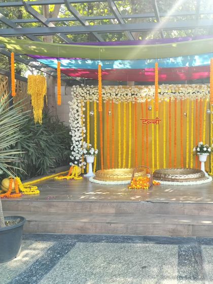 An outdoor Haldi setup on a wooden deck. The backdrop features yellow and orange drapes with strings of white and yellow flowers, and two brass urulis for the ceremony.