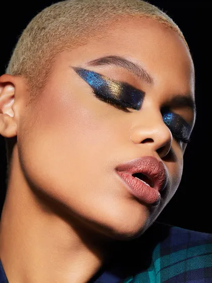 A dramatic, glittering winged eyeshadow look in shades of blue and gold on a model with a buzz cut, showcasing edgy and glamorous artistry.