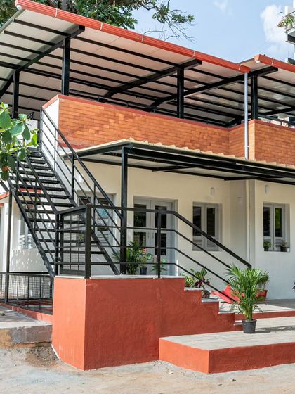 A wider view of the Anganwadi's exterior, showing the relationship between the ground floor, the staircase, and the semi-covered rooftop. The material palette of red-oxide, white plaster, and exposed brick is simple, cost-effective, and timeless.