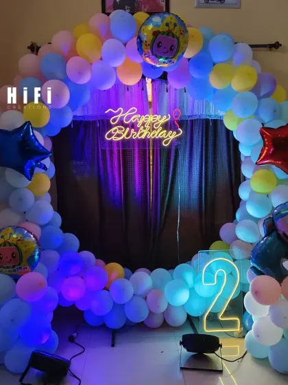 A Cocomelon-themed second birthday party setup for a home, featuring a circular balloon arch and neon sign.