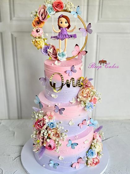 A magnificent three-tier cake for a magical first birthday. This pastel pink and purple creation is adorned with butterflies, flowers, and a custom fondant doll on a swing.