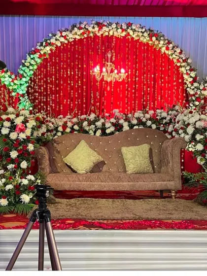 A classic reception stage with a rich red backdrop and a semi-circular arch of white and red flowers. The crystal chandelier adds a touch of glamour to the traditional design.