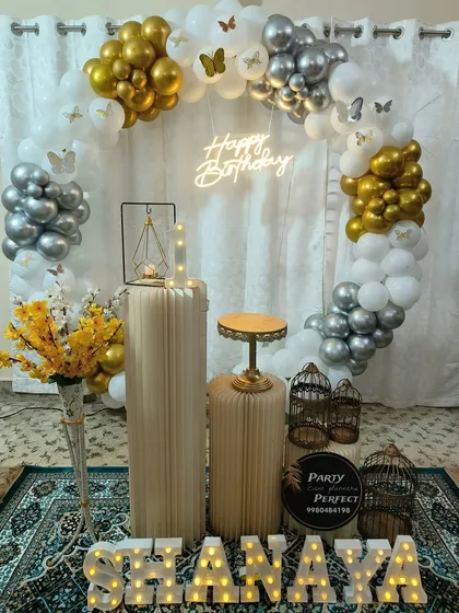 An elegant and airy birthday setup for Shanaya. The white, gold, and silver balloon ring is accented with butterflies and a neon sign, creating a light and celebratory feel.