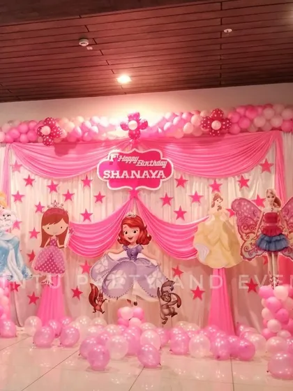 A princess themed first birthday stage decoration in pink. The setup features character cutouts, star details, and a full balloon arch over draped curtains.