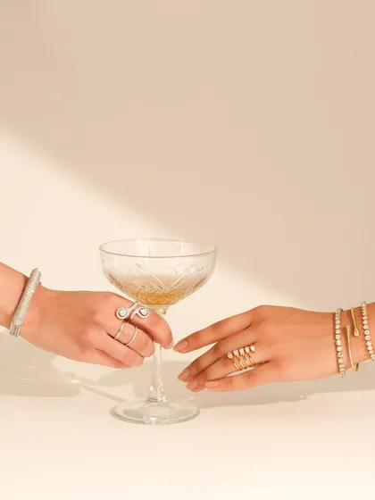 An elegant "Cocktail Edit" shot for a jewelry brand, featuring two hands adorned with diamond rings and bracelets reaching for a coupe glass. The image is sophisticated and aspirational.