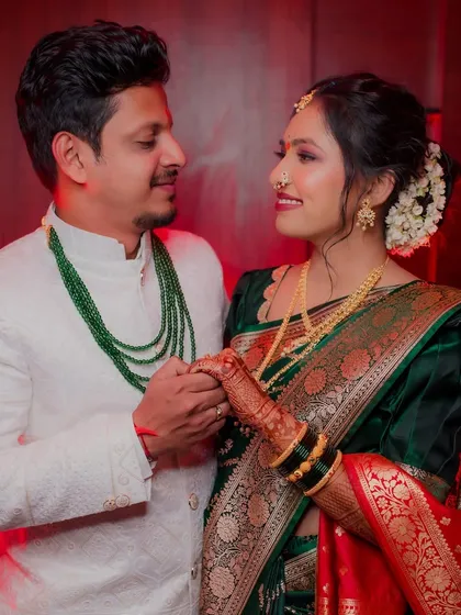 The couple holds hands and shares a loving gaze. This portrait highlights their traditional outfits, with the groom's emerald green necklace complementing the bride's beautiful saree.