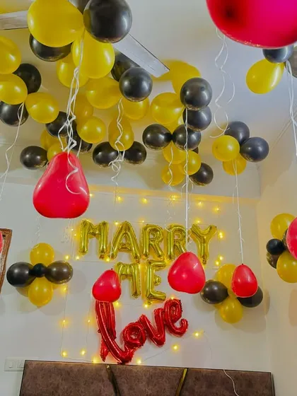A 'Marry Me' proposal setup in a room filled with yellow and black balloons, with fairy lights adding a warm glow.