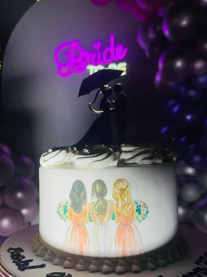 Details matter, especially for a bridal shower. This custom cake features a hand-painted illustration of the bride with her bridesmaids, topped with a silhouette of the couple. In the background, a "Bride to be" neon sign adds to the fun and celebratory atmosphere.