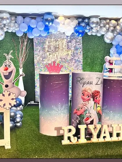 A Frozen theme party on an artificial grass floor. The setup includes a silver sequin wall, character cutouts, and a blue and silver balloon arch.