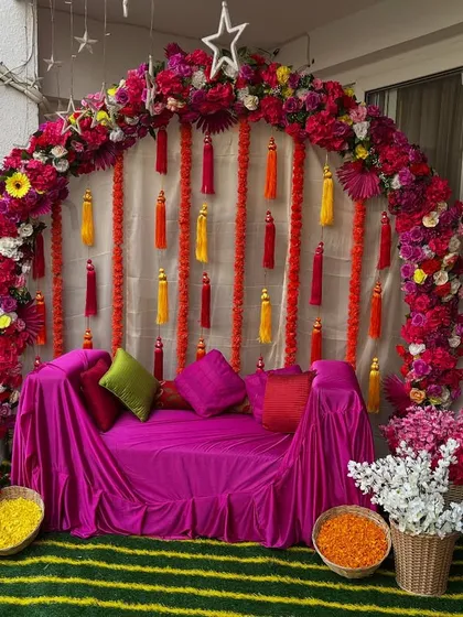 This vibrant mehndi seating at home features a semi-circular arch of bright pink and red flowers. The decor is complemented by colorful tassels and baskets of fresh flower petals.