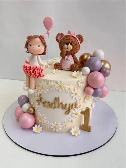 A full shot of the doll and teddy bear cake, personalized with the name "Aadhya" and the number "1" in gold.