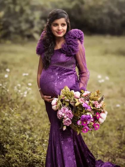 A stunning solo portrait in a field of wildflowers. The mother-to-be, in a dramatic purple gown, holds a large bouquet, looking like a queen of the meadow.
