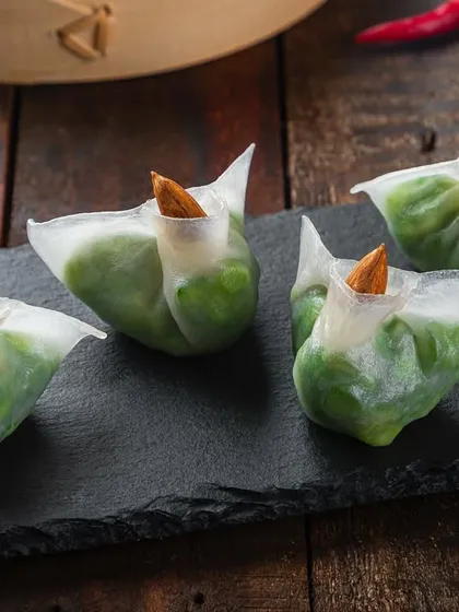 Translucent dumplings filled with green vegetables and topped with an almond. The macro focus on a dark slate platter showcases the intricate details.