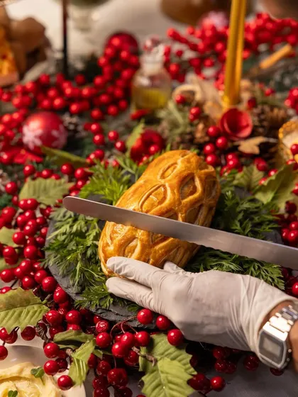 A chef's gloved hand carefully slices into the Lamb Wellington, revealing the perfectly cooked filling inside.