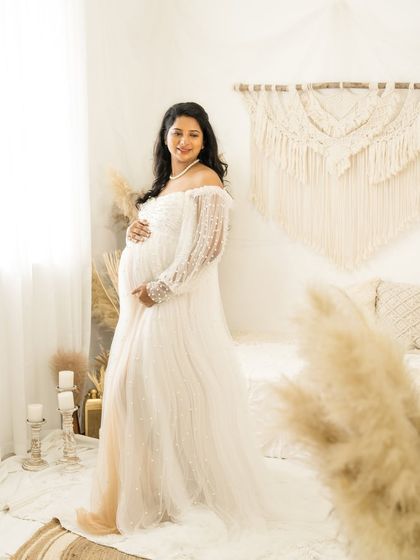 A graceful portrait of a mother-to-be in a white lace gown. She stands in a bright, boho-themed room, her hands on her bump, with a soft and happy expression.