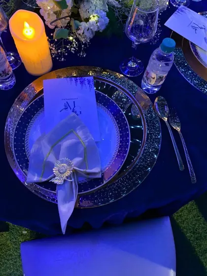 An opulent table setting under dramatic blue lighting. The combination of dark linens, ornate charger plates, and a bespoke napkin holder creates a luxurious and moody atmosphere for an exclusive evening event.
