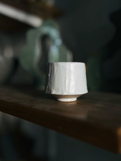 A small, faceted cup with a simple white glaze. The beauty is in the subtle, hand-carved shape that feels perfect to hold.