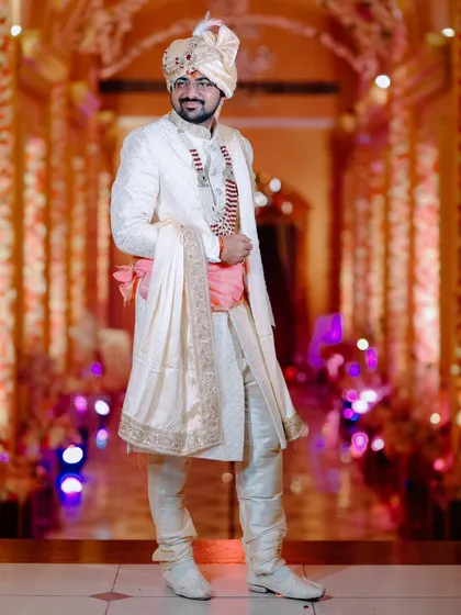 A full-length portrait of the groom in his elegant off-white sherwani. The warm, glowing background of the venue adds a touch of grandeur to his sharp look.