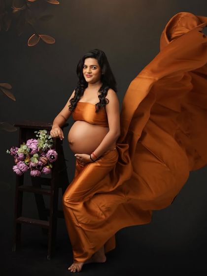A moment of pure elegance. This portrait captures the beauty of the baby bump with a flowing satin gown and a dramatic pose, set against a dark, moody backdrop for a fine art feel.