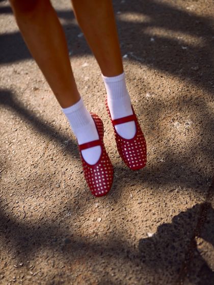 A sun-drenched shot of the red studded ballet flats. The light catches the silver studs, highlighting the craftsmanship and adding a dynamic quality to the vibrant red leather.