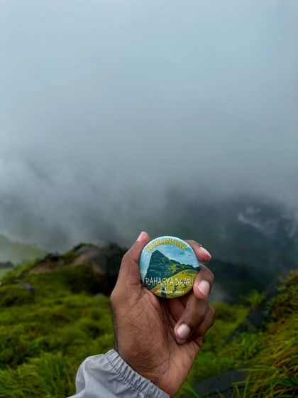 A close-up of our custom Kurinjal trek badge, held against the foggy, green landscape of the peak.