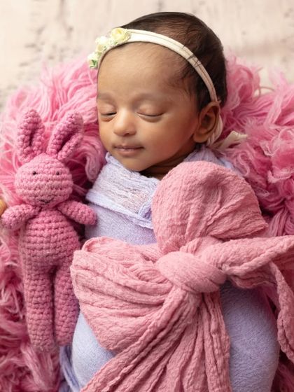 A tiny newborn baby wrapped in soft pink and purple fabrics, nestled in a fluffy basket with a little bunny friend.