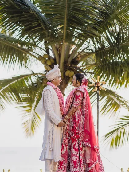 A beautiful couple shot against a backdrop of palm trees. The bride's classic makeup and red lehenga create a stunning contrast, making for a memorable wedding portrait.