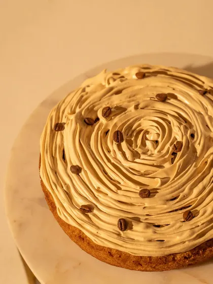 A close-up top-down view of a coffee tres leches cake, focusing on the swirl of the frosting and the sprinkled coffee beans. This detail shot emphasizes the dessert's texture and craftsmanship.