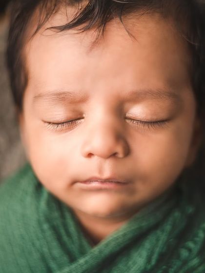 A close-up of a newborn's peaceful face, swaddled in a deep green wrap. The soft focus and gentle lighting highlight the serene beauty of this precious moment.