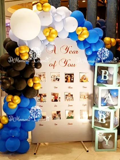 A "A Year of You" milestone photo board for the Boss Baby party. The display is framed by a blue, black, and white balloon arch, with "BABY" blocks alongside.