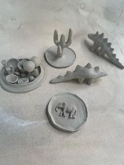 A collection of small, unfired sculptural pieces made in our hand-building course. You can see tiny dinosaurs, a cactus, a coral-themed dish, and a coaster with an elephant stamp.