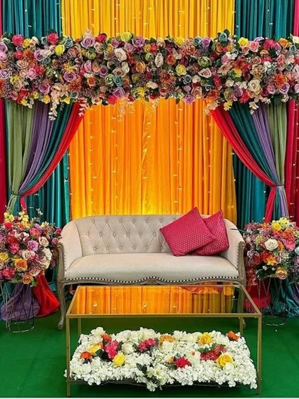 A vibrant and colorful Mehndi or Sangeet stage idea for a home function. The multi-colored drapes and lush floral arrangements create a rich and festive backdrop.