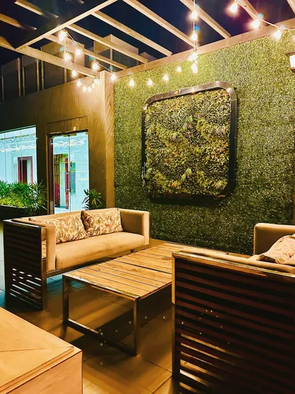 This cozy seating nook at a rooftop cafe is defined by a full-height artificial green wall and a framed living wall art piece. The warm string lights under the open-beam pergola create an intimate and relaxing atmosphere.