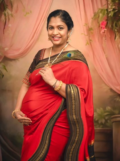 A beautiful solo portrait of a mom-to-be in a red saree, her hand gently on her bump, adorned with a peacock feather.