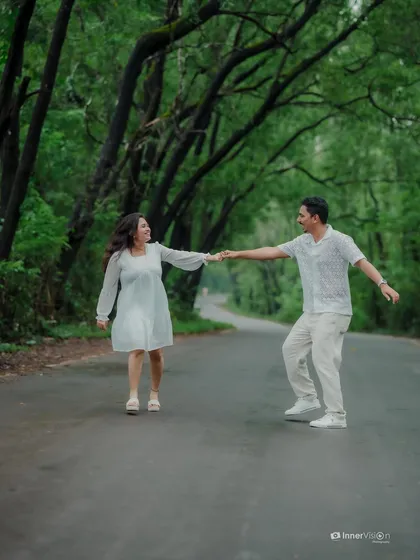 The couple takes a step in a dance-like pose on the road, holding hands and looking at each other. The lush green background makes their white outfits pop.