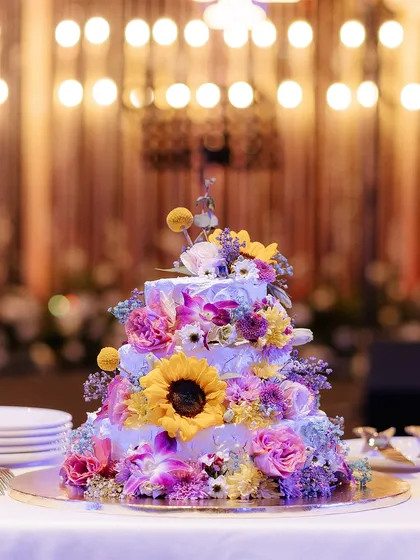 A stunning multi-tiered wedding cake, beautifully decorated with a cascade of fresh, colorful flowers including sunflowers and orchids.