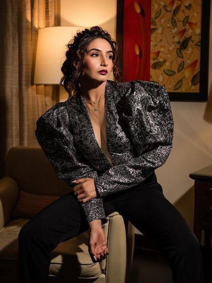 An indoor portrait of Ragini Dwivedi in a stylish silver and black top. The warm, soft lighting from the lamp creates an intimate and sophisticated mood.