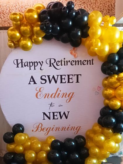 A balloon ring backdrop for a retirement party. The black and gold balloons frame a sign that reads 'Happy Retirement, A Sweet Ending to a New Beginning'.