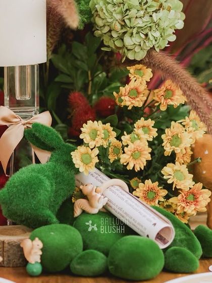 A close-up of a table centerpiece, featuring a mossy rabbit, a deer, and a scroll, all nestled among flowers.